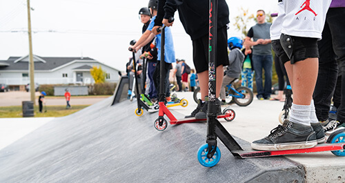 Kids with scooters are lined up at the top of a ramp at a skatepark.