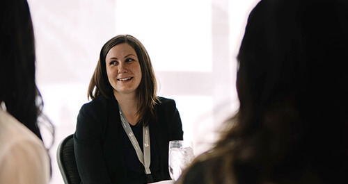 A woman is sitting at a table with two others, she is listening and smiling.